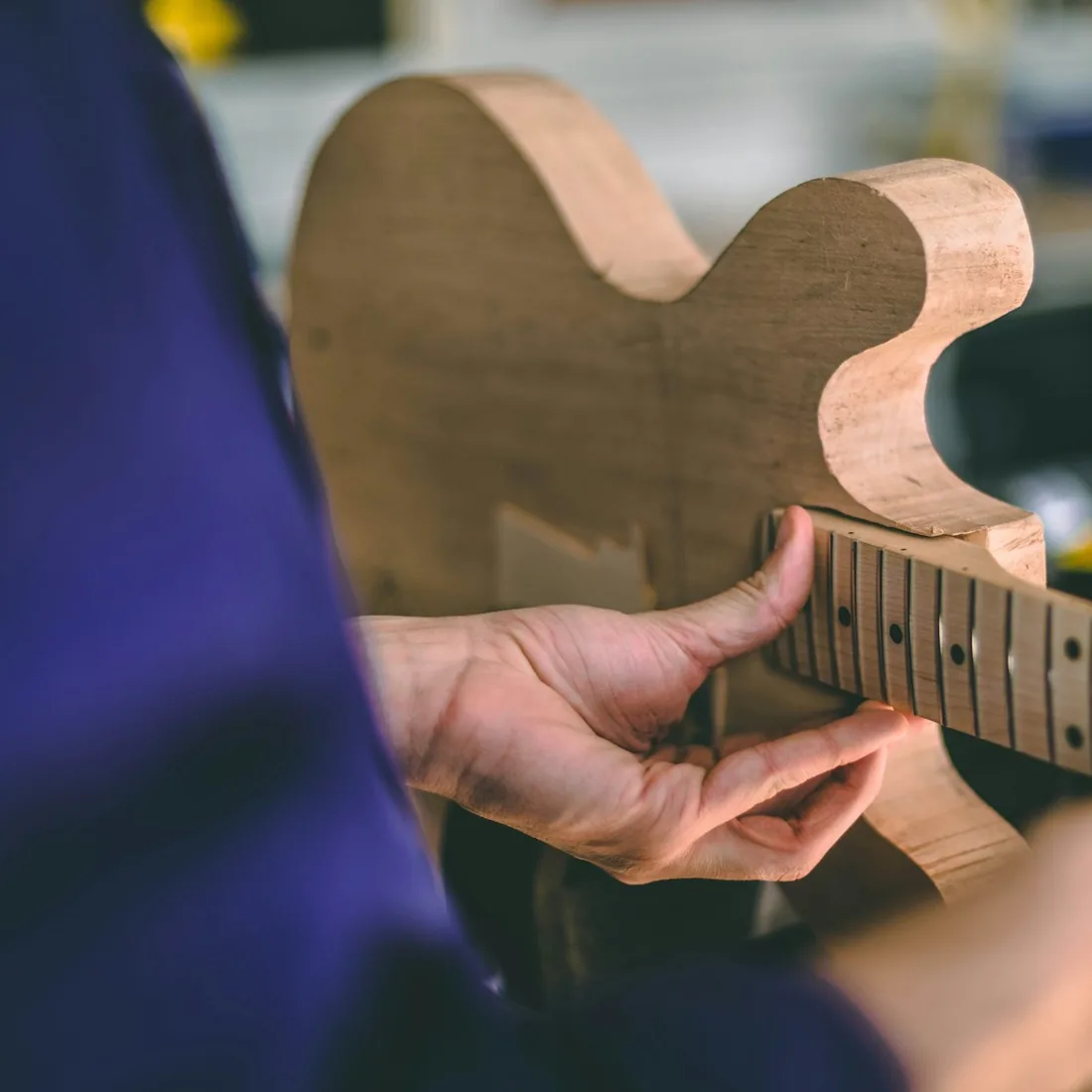 Manos ajustando el mástil y el diapasón sobre el cuerpo de una guitarra en madera clara, en un taller de luthería